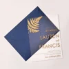 A sophisticated wedding invitation suite centered around a transparent acrylic invitation. The main card is partially tucked into a navy blue envelope with a shimmering metallic gold liner.