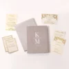 An elegant wedding invitation suite laid out on a clean white surface. The centerpiece is a textured, mauve-colored velvet booklet featuring white monogrammed letters separated by a horizontal line, with the date printed below. Surrounding the booklet are several cream-colored cards with Silver foil accents, including.