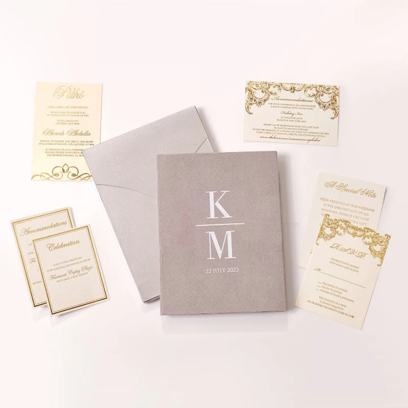 An elegant wedding invitation suite laid out on a clean white surface. The centerpiece is a textured, mauve-colored velvet booklet featuring white monogrammed letters separated by a horizontal line, with the date printed below. Surrounding the booklet are several cream-colored cards with Silver foil accents, including.