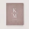 A top-down, centered shot of a portrait-oriented wedding invitation folder. The folder is covered in a dusty rose or mauve velvet fabric. In the center, white serif lettering displays a large separated by a horizontal line. Below the monogram, the date is printed in a smaller white font. The folder is set against a plain, light-colored background.