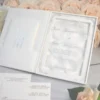 An open, luxury white folio wedding invitation set against a backdrop of peach and white roses. The right side of the folio features a clear acrylic invitation with silver foil lettering for "Blake Alexandra" and "Skyler Tate," resting on a bed of soft white petals. The left side has a pocket containing a secondary card with silver text. In the foreground, additional matching stationery is visible, including an RSVP card and a rehearsal dinner schedule. The entire suite is arranged on a light grey wood-textured surface next to a small decorative glass, creating a bright and sophisticated presentation.
