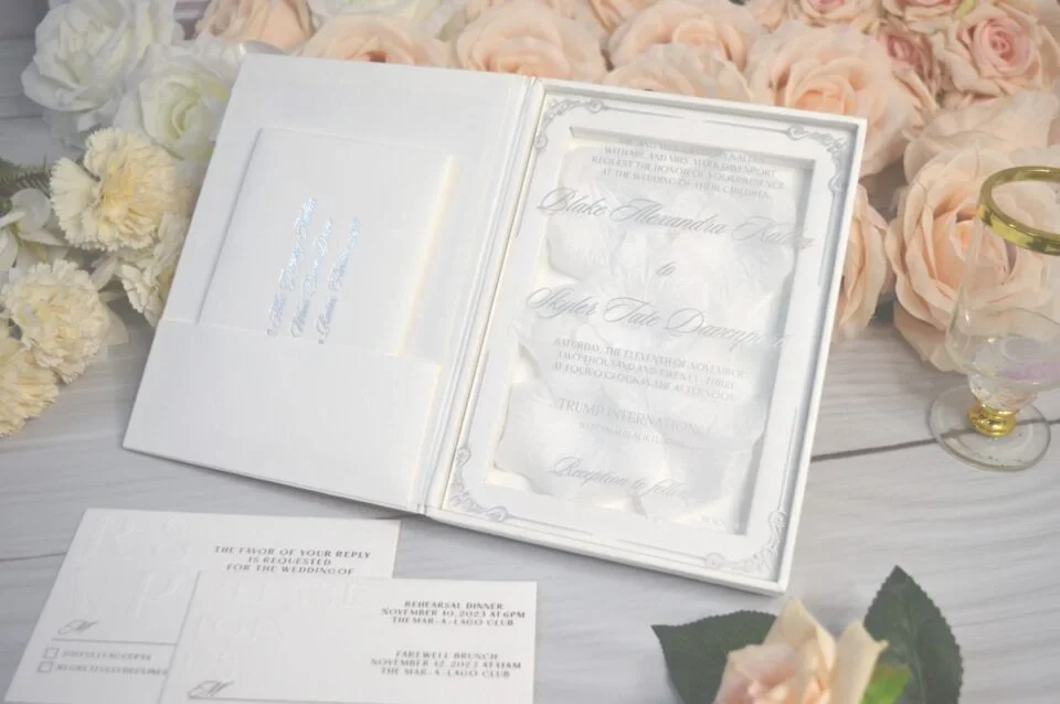 An open, luxury white folio wedding invitation set against a backdrop of peach and white roses. The right side of the folio features a clear acrylic invitation with silver foil lettering for "Blake Alexandra" and "Skyler Tate," resting on a bed of soft white petals. The left side has a pocket containing a secondary card with silver text. In the foreground, additional matching stationery is visible, including an RSVP card and a rehearsal dinner schedule. The entire suite is arranged on a light grey wood-textured surface next to a small decorative glass, creating a bright and sophisticated presentation.