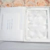 An open, luxury white folio wedding invitation set against a backdrop of peach and white roses. The right side of the folio features a clear acrylic invitation with silver foil lettering for "Blake Alexandra" and "Skyler Tate," resting on a bed of soft white petals. The left side has a pocket containing a secondary card with silver text. In the foreground, additional matching stationery is visible, including an RSVP card and a rehearsal dinner schedule. The entire suite is arranged on a light grey wood-textured surface next to a small decorative glass, creating a bright and sophisticated presentation.