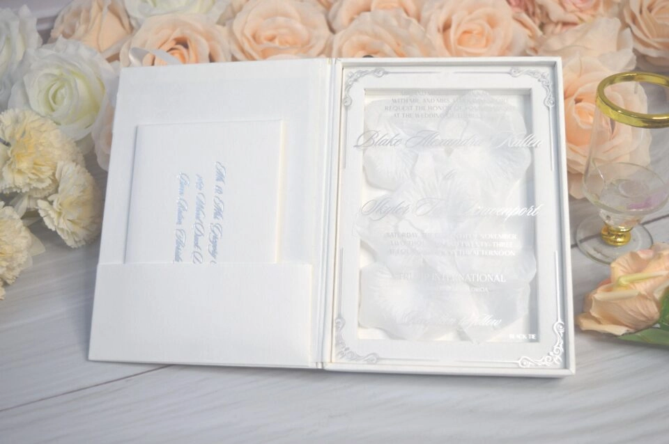 An open, luxury white folio wedding invitation set against a backdrop of peach and white roses. The right side of the folio features a clear acrylic invitation with silver foil lettering for "Blake Alexandra" and "Skyler Tate," resting on a bed of soft white petals. The left side has a pocket containing a secondary card with silver text. In the foreground, additional matching stationery is visible, including an RSVP card and a rehearsal dinner schedule. The entire suite is arranged on a light grey wood-textured surface next to a small decorative glass, creating a bright and sophisticated presentation.