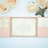 A high-angle view of an open silk gatefold invitation folio displayed against a clean white background, surrounded by several peach-colored roses and a small bronze Eiffel Tower figurine.The elegant wedding suite includes:The center panel: A formal white invitation card with gold-foiled text and an ornate scrollwork border.It announces the marriage of Stephanie Elaine Godoy and William Benjamin Schilling Side pockets: The left and right panels feature pockets holding coordinating stationery. On the left, two small cards with gold monograms are visible; on the right, a larger enclosure card is tucked away.The design: The folio is crafted from textured peach silk, with the main card secured in place by four matching silk corner tabs. The soft color palette and metallic gold accents create a romantic, high-end aesthetic for a formal wedding celebration.