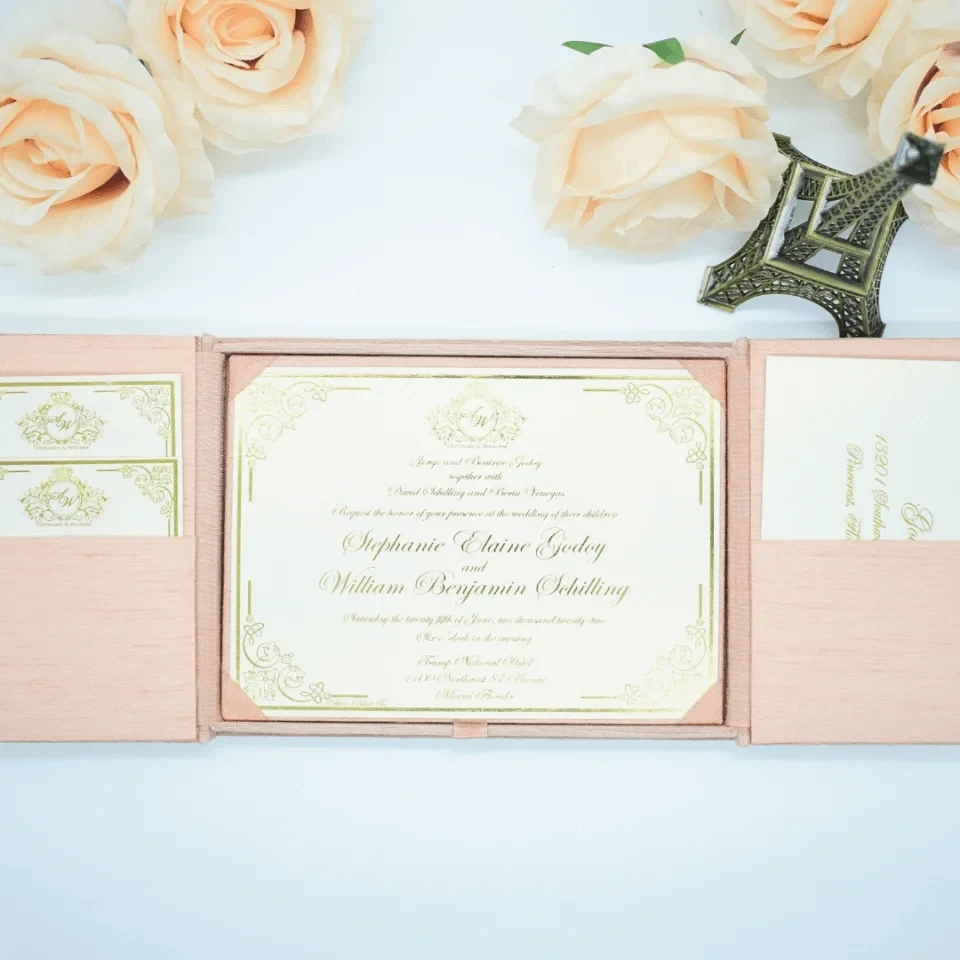 A high-angle view of an open silk gatefold invitation folio displayed against a clean white background, surrounded by several peach-colored roses and a small bronze Eiffel Tower figurine.The elegant wedding suite includes:The center panel: A formal white invitation card with gold-foiled text and an ornate scrollwork border.It announces the marriage of Stephanie Elaine Godoy and William Benjamin Schilling Side pockets: The left and right panels feature pockets holding coordinating stationery. On the left, two small cards with gold monograms are visible; on the right, a larger enclosure card is tucked away.The design: The folio is crafted from textured peach silk, with the main card secured in place by four matching silk corner tabs. The soft color palette and metallic gold accents create a romantic, high-end aesthetic for a formal wedding celebration.