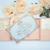 A high-angle view of an open silk gatefold invitation folio displayed against a clean white background, surrounded by several peach-colored roses and a small bronze Eiffel Tower figurine.The elegant wedding suite includes:The center panel: A formal white invitation card with gold-foiled text and an ornate scrollwork border.It announces the marriage of Stephanie Elaine Godoy and William Benjamin Schilling Side pockets: The left and right panels feature pockets holding coordinating stationery. On the left, two small cards with gold monograms are visible; on the right, a larger enclosure card is tucked away.The design: The folio is crafted from textured peach silk, with the main card secured in place by four matching silk corner tabs. The soft color palette and metallic gold accents create a romantic, high-end aesthetic for a formal wedding celebration.