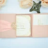 A high-angle view of an open silk gatefold invitation folio displayed against a clean white background, surrounded by several peach-colored roses and a small bronze Eiffel Tower figurine.The elegant wedding suite includes:The center panel: A formal white invitation card with gold-foiled text and an ornate scrollwork border.It announces the marriage of Stephanie Elaine Godoy and William Benjamin Schilling Side pockets: The left and right panels feature pockets holding coordinating stationery. On the left, two small cards with gold monograms are visible; on the right, a larger enclosure card is tucked away.The design: The folio is crafted from textured peach silk, with the main card secured in place by four matching silk corner tabs. The soft color palette and metallic gold accents create a romantic, high-end aesthetic for a formal wedding celebration.