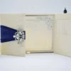 An open, cream-colored gatefold wedding invitation suite displayed on a white surface. The central panel features the formal invitation for "Allen & Maria," printed in elegant black script and serif fonts with a delicate floral border in the corners. The two side flaps contain pockets holding additional information cards. Above the invitation, three decorative roses—two white and one pale peach—are arranged alongside a small bronze Eiffel Tower figurine. The scene is brightly lit, emphasizing the textured fabric of the invitation holder and the clean, romantic aesthetic.