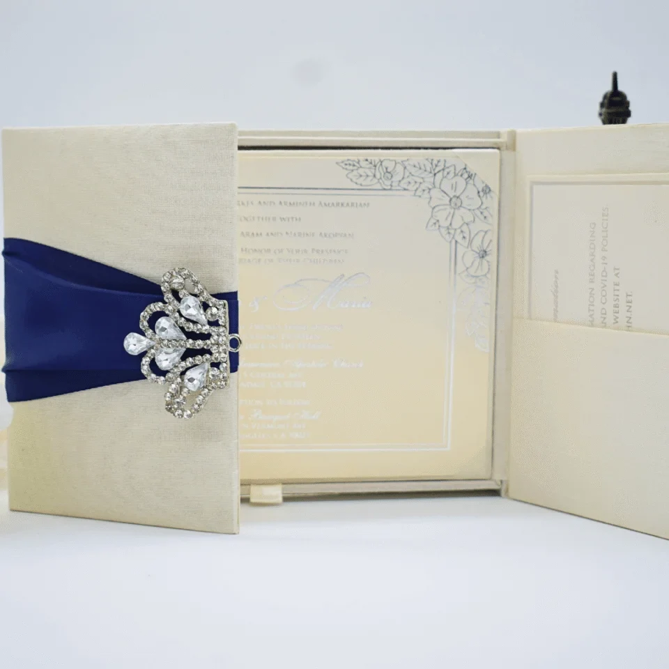 An open, cream-colored gatefold wedding invitation suite displayed on a white surface. The central panel features the formal invitation for "Allen & Maria," printed in elegant black script and serif fonts with a delicate floral border in the corners. The two side flaps contain pockets holding additional information cards. Above the invitation, three decorative roses—two white and one pale peach—are arranged alongside a small bronze Eiffel Tower figurine. The scene is brightly lit, emphasizing the textured fabric of the invitation holder and the clean, romantic aesthetic.