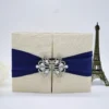 A cream-colored, silk-textured gatefold invitation box sits centered on a white surface. A wide, royal blue satin ribbon wraps horizontally around the middle, gathered at the center by a large, ornate silver brooch encrusted with clear crystals in a symmetrical, butterfly-like pattern. In the background, a small bronze Eiffel Tower figurine and the soft edges of a pale peach flower are visible, adding a decorative touch to the flat lay composition.