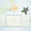 An open, cream-colored gatefold wedding invitation suite displayed on a white surface. The central panel features the formal invitation for "Allen & Maria," printed in elegant black script and serif fonts with a delicate floral border in the corners. The two side flaps contain pockets holding additional information cards. Above the invitation, three decorative roses—two white and one pale peach—are arranged alongside a small bronze Eiffel Tower figurine. The scene is brightly lit, emphasizing the textured fabric of the invitation holder and the clean, romantic aesthetic.