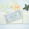 An open, cream-colored gatefold wedding invitation suite displayed on a white surface. The central panel features the formal invitation for "Allen & Maria," printed in elegant black script and serif fonts with a delicate floral border in the corners. The two side flaps contain pockets holding additional information cards. Above the invitation, three decorative roses—two white and one pale peach—are arranged alongside a small bronze Eiffel Tower figurine. The scene is brightly lit, emphasizing the textured fabric of the invitation holder and the clean, romantic aesthetic.