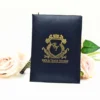 A top-down view of a sophisticated black silk wedding invitation folder on a white surface. The cover features a striking gold-foil crest in the center, topped with a crown and containing the monogrammed initials Below the crest, the names are printed in bold, gold-foil capital letters.