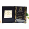 An elegant, black and gold wedding invitation suite displayed on a white surface, surrounded by peach and white roses and a small Eiffel Tower figurine. The main invitation is housed in a black velvet-style folder. On the right side, the invitation is printed on a clear acrylic or glossy black material with ornate gold filigree borders. The text, also in gold, announces the wedding.