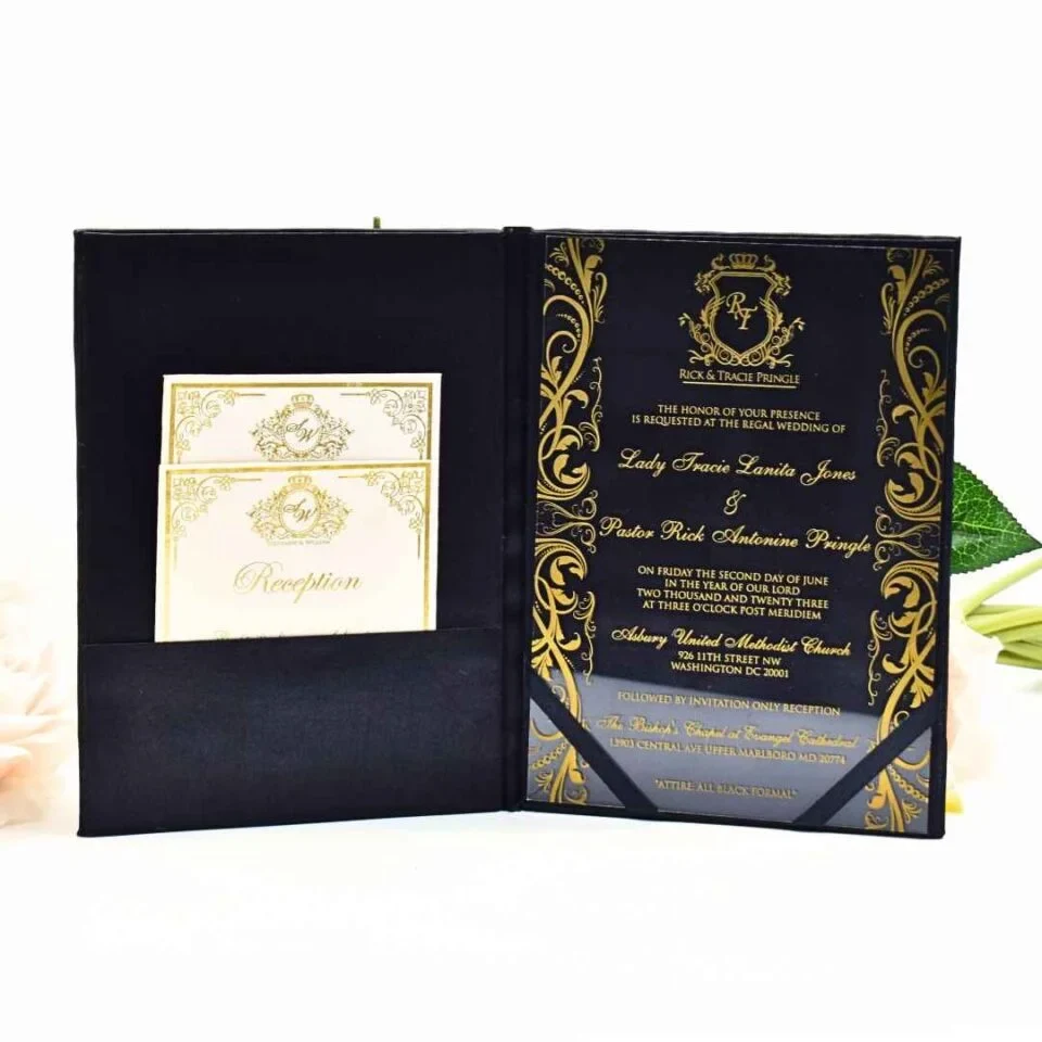 An elegant, black and gold wedding invitation suite displayed on a white surface, surrounded by peach and white roses and a small Eiffel Tower figurine. The main invitation is housed in a black velvet-style folder. On the right side, the invitation is printed on a clear acrylic or glossy black material with ornate gold filigree borders. The text, also in gold, announces the wedding.
