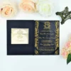 An elegant, black and gold wedding invitation suite displayed on a white surface, surrounded by peach and white roses and a small Eiffel Tower figurine. The main invitation is housed in a black velvet-style folder. On the right side, the invitation is printed on a clear acrylic or glossy black material with ornate gold filigree borders. The text, also in gold, announces the wedding.