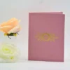 A sophisticated dusty rose or mauve wedding invitation folder** standing vertically on a white surface, flanked by soft peach and white roses. The front of the folder features a luxurious **gold foil-stamped crest** in the center. The crest includes the initials encircled by an ornate wreath and flanked by intricate floral and vine-like gold patterns. The folder is slightly open, suggesting a book-style or gatefold design, and the lighting highlights the subtle texture of the rose-colored fabric or cardstock.