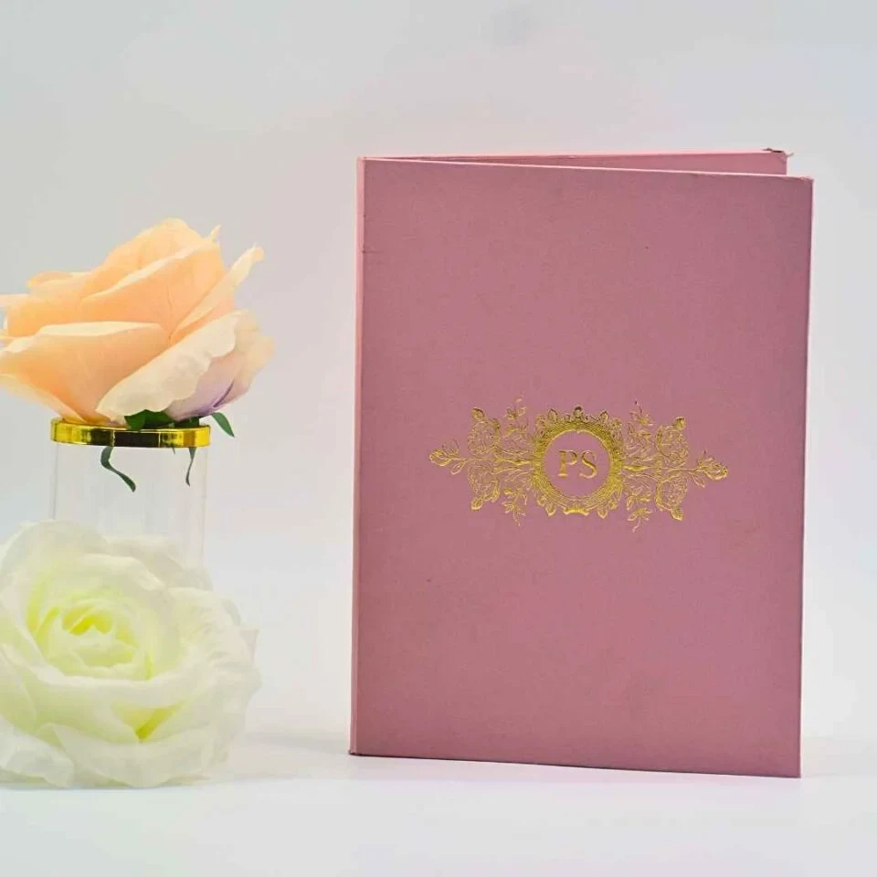 A sophisticated dusty rose or mauve wedding invitation folder** standing vertically on a white surface, flanked by soft peach and white roses. The front of the folder features a luxurious **gold foil-stamped crest** in the center. The crest includes the initials encircled by an ornate wreath and flanked by intricate floral and vine-like gold patterns. The folder is slightly open, suggesting a book-style or gatefold design, and the lighting highlights the subtle texture of the rose-colored fabric or cardstock.