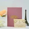 A dusty rose wedding invitation suite displayed on a white surface, featuring a vertical folder, a separate RSVP card, and decorative accents. The mauve-colored folder stands in the background, adorned with a central gold foil-stamped crest containing the initials surrounded by floral and filigree patterns. Propped up in front of the folder is a cream-colored RSVP card featuring matching gold foil scrollwork and a small fleur-de-lis symbol. The card includes response instructions and a wedding website link. To the left of the stationery is a peach-colored rose in a glass vase with a gold rim, and to the right is a small bronze Eiffel Tower figurine, creating a romantic and elegant setting.