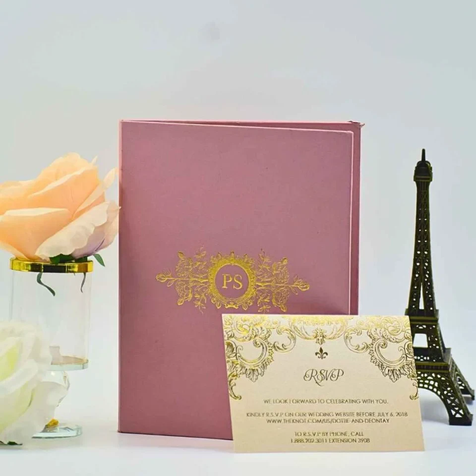 A dusty rose wedding invitation suite displayed on a white surface, featuring a vertical folder, a separate RSVP card, and decorative accents. The mauve-colored folder stands in the background, adorned with a central gold foil-stamped crest containing the initials surrounded by floral and filigree patterns. Propped up in front of the folder is a cream-colored RSVP card featuring matching gold foil scrollwork and a small fleur-de-lis symbol. The card includes response instructions and a wedding website link. To the left of the stationery is a peach-colored rose in a glass vase with a gold rim, and to the right is a small bronze Eiffel Tower figurine, creating a romantic and elegant setting.