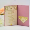 An open dusty rose wedding invitation folder standing upright on a white surface, displaying the full invitation suite alongside a peach rose and a separate RSVP card. Left Panel: Contains the main invitation card, which is cream-colored with metallic gold foil text and intricate scrollwork. It features a prominent fleur-de-lis and announces the wedding.