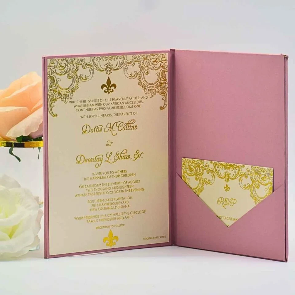 An open dusty rose wedding invitation folder standing upright on a white surface, displaying the full invitation suite alongside a peach rose and a separate RSVP card. Left Panel: Contains the main invitation card, which is cream-colored with metallic gold foil text and intricate scrollwork. It features a prominent fleur-de-lis and announces the wedding.