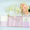 A high-angle view of an open pink silk gatefold invitation folio displayed on a white surface, accompanied by white and peach roses. The fairy-tale-themed suite includes: The center panel: Features a pink invitation card with a gold-bordered frame and the heading. It announces the celebration for in elegant gold script. Side pockets: The left and right panels contain pockets holding coordinating white cards with gold-foiled accents.Decorative details: Intricate gold laser-cut butterflies are tucked into the corners of the main invitation, which is secured by a thin pink satin ribbon pull-tab. The overall design is whimsical and romantic, using a soft pastel palette and shimmering gold highlights.