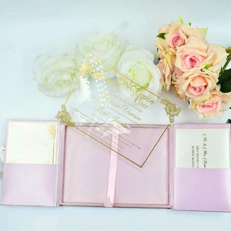 A high-angle view of an open pink silk gatefold invitation folio displayed on a white surface, accompanied by white and peach roses. The fairy-tale-themed suite includes: The center panel: Features a pink invitation card with a gold-bordered frame and the heading. It announces the celebration for in elegant gold script. Side pockets: The left and right panels contain pockets holding coordinating white cards with gold-foiled accents.Decorative details: Intricate gold laser-cut butterflies are tucked into the corners of the main invitation, which is secured by a thin pink satin ribbon pull-tab. The overall design is whimsical and romantic, using a soft pastel palette and shimmering gold highlights.