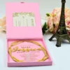 A high-angle view of a luxurious, open emerald velvet wedding invitation box displayed on a light surface. The stationery suite includes:Inside Lid: A gold-bordered card featuring an elegant gold-etched illustration of a historic, multi-story building with palm trees. The Base:A clear acrylic invitation card rests in a recessed compartment. It features the names and wedding details in a mix of white script and serif typography.Accessories: A thick emerald green silk tassel is attached to the invitation, and a matching green velvet band with a gold wax seal is visible in the base.Decor: A small bronze Eiffel Tower figurine stands to the right, and a soft peach-colored rose is positioned on the left, adding to the sophisticated travel-themed aesthetic.