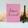 A front-facing view of a vertical, rectangular blush pink invitation box standing upright against a white background. The lid is personalized with the names printed in a large, elegant gold foil script that spans the center of the box. The presentation is styled with decorative accents: A small, dark bronze Eiffel Tower figurine** stands to the right. Large, soft peach and cream-colored roses** are arranged behind and to the sides of the box, creating a romantic frame. The clean design, combined with the vibrant pink hue and shimmering gold typography, offers a modern and luxurious look for a wedding or special event.