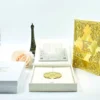 An open, **luxurious cream-colored wedding invitation box** showcasing a multi-piece stationery suite. The arrangement includes: * An **inner lid** featuring a laser-cut silver pocket holding an RSVP card. * A stunning **gold mirrored acrylic invitation card** in the foreground, etched with white calligraphy for "Maria Goretti Numfor and Oscar C. Ambe." The card is embellished with small raised gold studs. * A **cream silk folio** with a central band tucked inside the box. * Decorative elements in the background, including a small **bronze Eiffel Tower**, white and peach roses, and a gold filigree-topped box. * The entire set is presented on a clean, bright white surface, highlighting the reflective gold and soft cream textures.