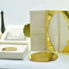 An open, **luxurious cream-colored wedding invitation box** showcasing a multi-piece stationery suite. The arrangement includes: * An **inner lid** featuring a laser-cut silver pocket holding an RSVP card. * A stunning **gold mirrored acrylic invitation card** in the foreground, etched with white calligraphy for "Maria Goretti Numfor and Oscar C. Ambe." The card is embellished with small raised gold studs. * A **cream silk folio** with a central band tucked inside the box. * Decorative elements in the background, including a small **bronze Eiffel Tower**, white and peach roses, and a gold filigree-topped box. * The entire set is presented on a clean, bright white surface, highlighting the reflective gold and soft cream textures.