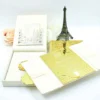 An open, **luxurious cream-colored wedding invitation box** showcasing a multi-piece stationery suite. The arrangement includes: * An **inner lid** featuring a laser-cut silver pocket holding an RSVP card. * A stunning **gold mirrored acrylic invitation card** in the foreground, etched with white calligraphy for "Maria Goretti Numfor and Oscar C. Ambe." The card is embellished with small raised gold studs. * A **cream silk folio** with a central band tucked inside the box. * Decorative elements in the background, including a small **bronze Eiffel Tower**, white and peach roses, and a gold filigree-topped box. * The entire set is presented on a clean, bright white surface, highlighting the reflective gold and soft cream textures.