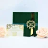 A high-angle shot of a luxurious wedding invitation suite. An open **emerald green velvet folio** features a reflective **gold mirror acrylic invitation** with white script and a matching monogram crest. In the foreground, a white **RSVP card** with intricate gold floral patterns and a QR code lies flat. The arrangement is accented by two soft **pale peach roses** on the right, all set against a clean, light blue background.