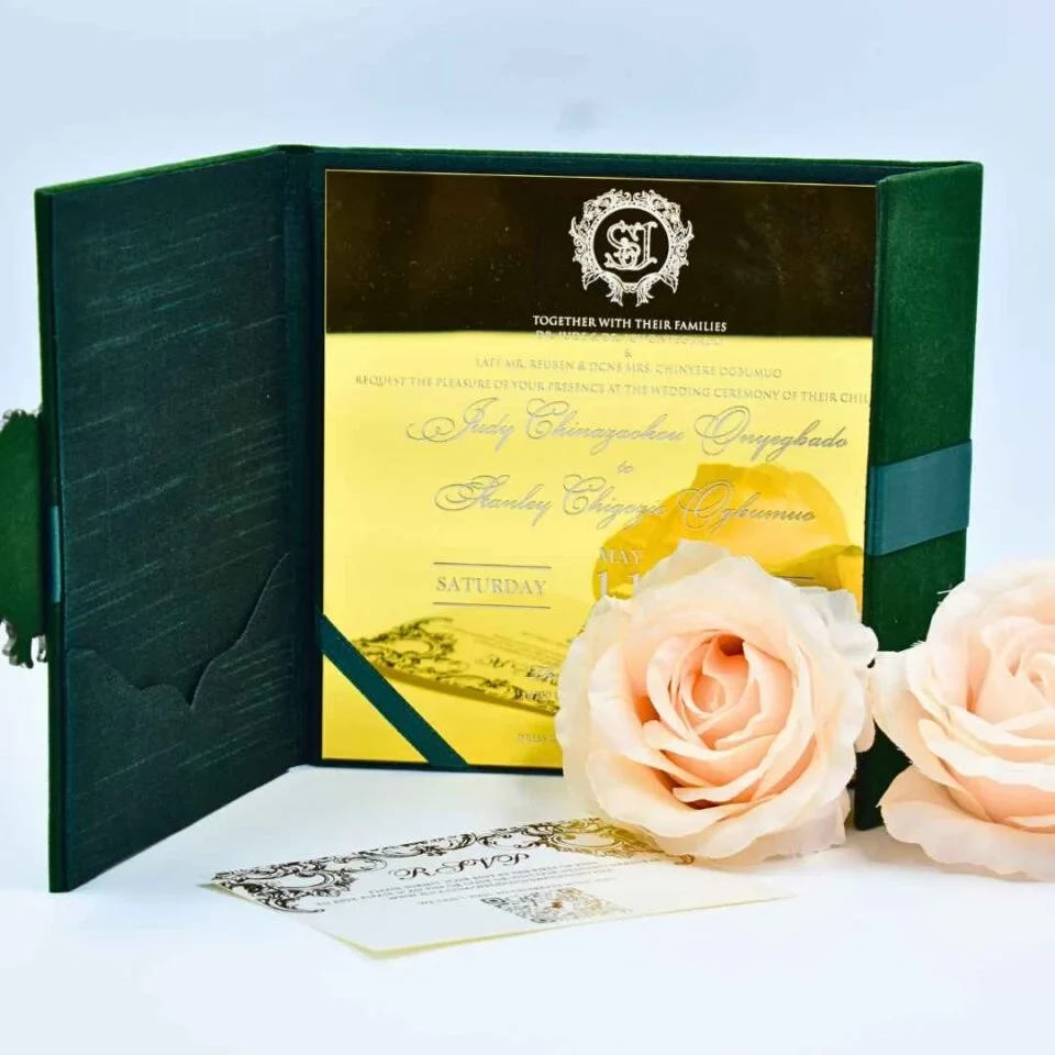 An open **emerald green velvet folio** displaying a wedding invitation for The main invitation is printed on a reflective **gold mirror acrylic** card with a white crest and script. The left side of the folio features a pocket, and an **RSVP card** with gold flourishes lies on the surface in front. Two large, **pale peach roses** are positioned in the foreground, creating a romantic and luxurious aesthetic against the light blue background.