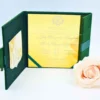 An open **emerald green velvet folio** displaying a wedding invitation for The invitation is printed on a highly reflective **gold mirror acrylic** card with elegant white script. The left side of the folio features a pocket containing a white RSVP card with yellow-gold floral details. A single **pale peach rose** is positioned in the foreground on the right, set against a clean, light blue background.