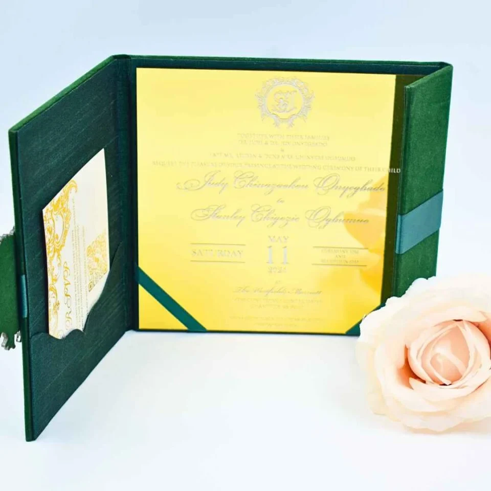 An open **emerald green velvet folio** displaying a wedding invitation for The invitation is printed on a highly reflective **gold mirror acrylic** card with elegant white script. The left side of the folio features a pocket containing a white RSVP card with yellow-gold floral details. A single **pale peach rose** is positioned in the foreground on the right, set against a clean, light blue background.