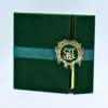 A square, luxurious **emerald green velvet gatefold folio** is shown closed. It is wrapped with a matching green satin ribbon and secured in the center by a large, ornate **gold metallic crest**. The crest features an intricate, interlocking monogram design. A sliver of the highly reflective **gold mirror acrylic invitation** inside is visible through the gatefold opening. The entire suite is set against a clean, light blue background.