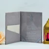 An elegant wedding invitation suite featuring a **Gray velvet folio** shown closed. The front of the folio is minimalist and modern, featuring the initials in a clean white serif font, separated by a horizontal line, with the date printed below. To the left of the stationery, a single, lush **pale peach rose** adds a soft, romantic touch. The entire suite is set against a clean, light blue background with subtle shadowing.