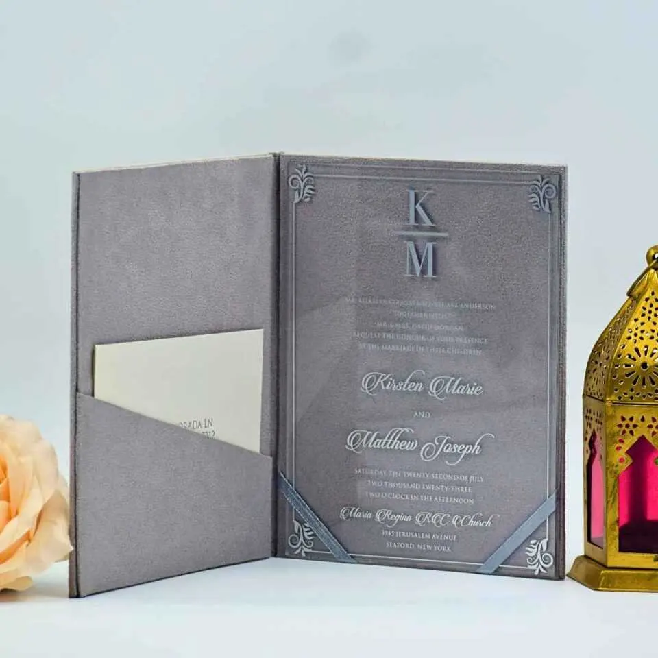 An elegant wedding invitation suite featuring a **Gray velvet folio** shown closed. The front of the folio is minimalist and modern, featuring the initials in a clean white serif font, separated by a horizontal line, with the date printed below. To the left of the stationery, a single, lush **pale peach rose** adds a soft, romantic touch. The entire suite is set against a clean, light blue background with subtle shadowing.