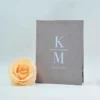 An open **Grey velvet folio** displays a wedding invitation for **"Kirsten Marie and Matthew Joseph."** The invitation is printed on a **clear acrylic card** featuring white script, a central monogram, and silver decorative corner flourishes. In the foreground to the left, a matching cream-colored envelope addressed to sits flat. The entire suite is photographed against a clean, light blue background with a hint of a floral element visible on the far left.