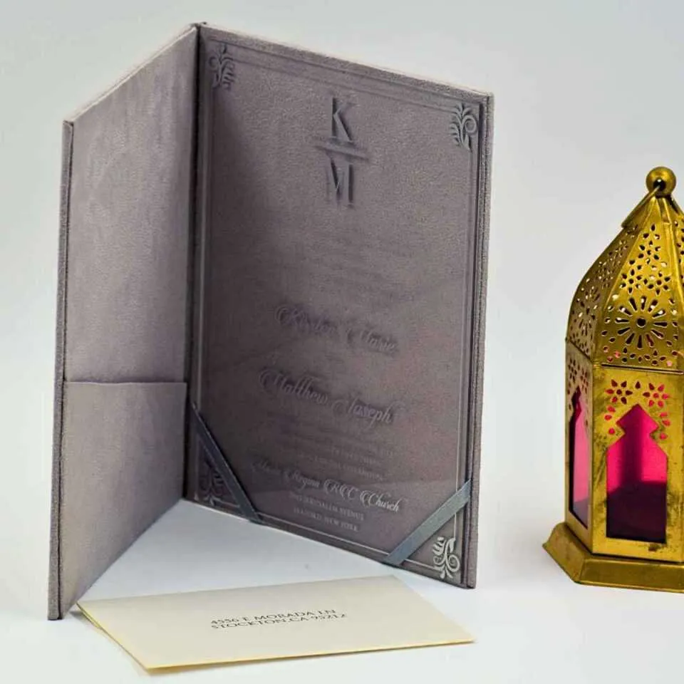 An open **Grey velvet folio** displays a wedding invitation for **"Kirsten Marie and Matthew Joseph."** The invitation is printed on a **clear acrylic card** featuring white script, a central monogram, and silver decorative corner flourishes. In the foreground to the left, a matching cream-colored envelope addressed to sits flat. The entire suite is photographed against a clean, light blue background with a hint of a floral element visible on the far left.