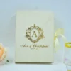 An elegant, ivory-colored invitation box or guest book featuring gold foil lettering. In the center is a large, stylized gold crest with the letter "A" inside an oval frame. Below the crest, the name written in a sophisticated gold script, followed by the date. A pale ivory ribbon is tied on the right side, and a soft peach-colored rose is partially visible in the bottom-left corner.