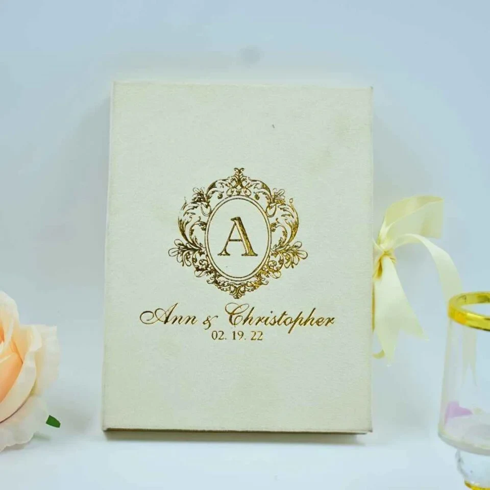 An elegant, ivory-colored invitation box or guest book featuring gold foil lettering. In the center is a large, stylized gold crest with the letter "A" inside an oval frame. Below the crest, the name written in a sophisticated gold script, followed by the date. A pale ivory ribbon is tied on the right side, and a soft peach-colored rose is partially visible in the bottom-left corner.