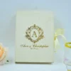 An elegant, ivory-colored invitation box or guest book featuring gold foil lettering. In the center is a large, stylized gold crest with the letter "A" inside an oval frame. Below the crest, the name written in a sophisticated gold script, followed by the date. A pale ivory ribbon is tied on the right side, and a soft peach-colored rose is partially visible in the bottom-left corner.