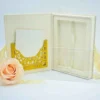 An open, luxurious cream-colored wedding invitation folio On the left panel, a white card sits inside an intricate, gold laser-cut pocket with floral scrollwork. The right panel features a deep recessed frame designed to hold the invitation. Leaning against the front is a clear acrylic invitation card with elegant white script. A soft peach rose and a pale yellow ribbon decorate the foreground against a clean white background.