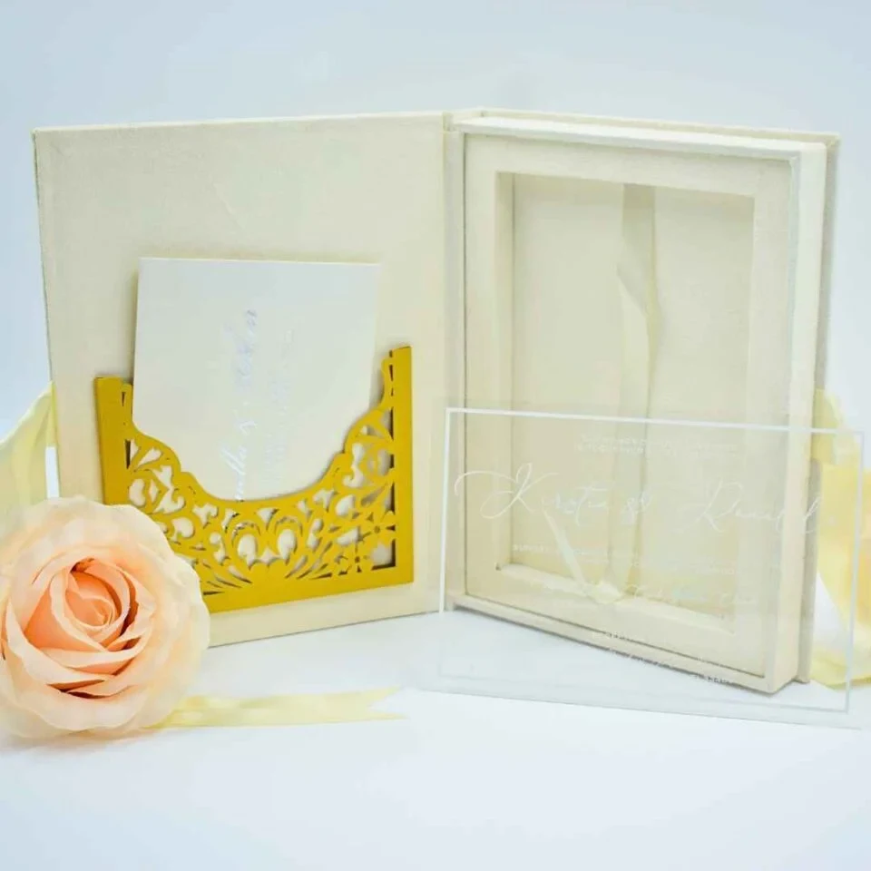An open, luxurious cream-colored wedding invitation folio On the left panel, a white card sits inside an intricate, gold laser-cut pocket with floral scrollwork. The right panel features a deep recessed frame designed to hold the invitation. Leaning against the front is a clear acrylic invitation card with elegant white script. A soft peach rose and a pale yellow ribbon decorate the foreground against a clean white background.