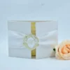 An elegant, closed **off-white wedding invitation folder** standing upright against a clean, light blue-grey background. The folder has a subtle, linen-like texture and is held shut by a wide, **white satin belly band**. At the center of the band is a decorative oval crest with a **gold-foil filigree border** and a scripted "B" monogram in the middle. The folder is slightly open, revealing a gold-patterned card inside. To the right, a large, soft **peach-colored rose** is partially visible in the foreground, adding a romantic touch to the composition.
