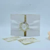 A professional product shot of a white textured wedding invitation folder standing upright, held closed by a wide white satin belly band. The center of the band features an ornate, oval-shaped crest with a gold-foil filigree border and a scripted "B" monogram. The folder is slightly open, showing a hint of the gold-foil wedding invitation inside. In the foreground, two coordinating inserts—an RSVP card and an Additional Details card—are laid flat, displaying matching gold baroque designs and QR codes. A soft peach rose is partially visible on the right edge, all set against a clean, neutral background.